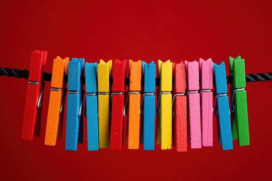 Row of colorful wooden clothespins clipped on black rope. Clothes pegs in bright red blue yellow pink orange and green arranged neatly on dark cord. Simple household objects for laundry or crafts.