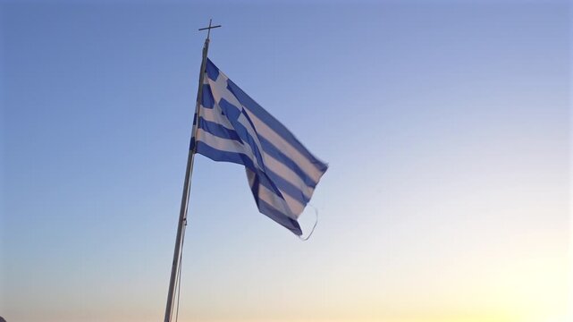 Greek flag waving in the sea breeze at sunset, with the blue and white national flag moving against a clear evening sky in warm Mediterranean light.