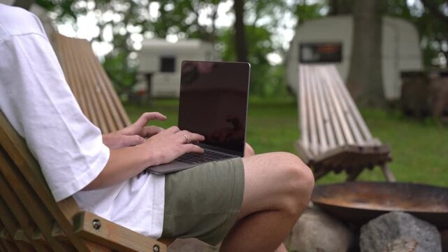 Person sits in wooden chair, typing on laptop outdoors. Grass and trees surround relaxed camper. Scene suggests leisurely work or vacation. Ideal for remote work, travel, or camping content