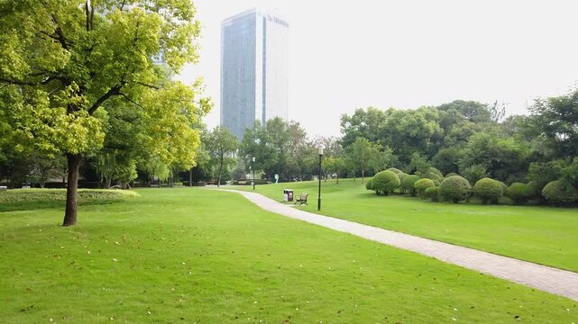 A well-maintained urban park with green lawn, trees, pathway, and a distant high-rise building under bright daylight.