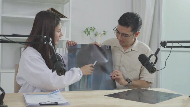 Asian female doctor explaining a chest X-ray film to a male host during a medical podcast interview. Recording a healthcare audio broadcast in a clinic studio. Respiratory health concept.