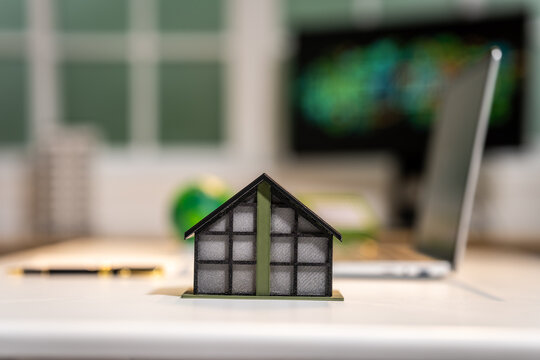 Small house model on a desk with a blurred laptop in the background, showing home, property, or real estate planning in an office setting