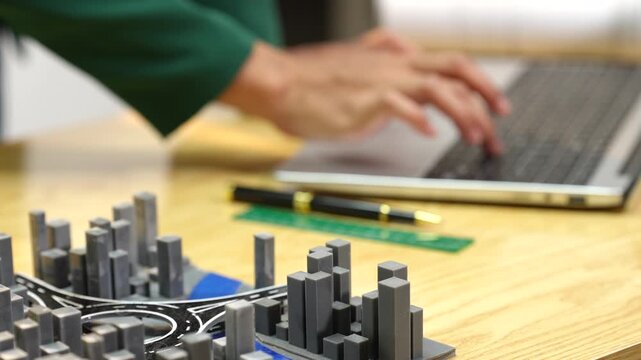 Blurred person working on a laptop beside a city model and drafting tools on a desk, showing urban planning and design work