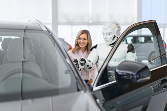 A humanoid robot stands next to a smiling woman, gesturing towards a car in a modern dealership showroom