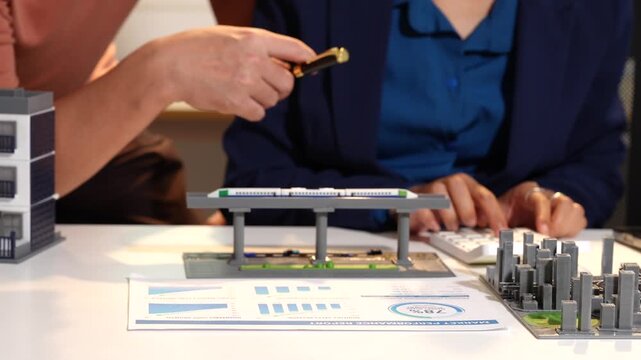 Business people discussing an architectural scale model during a planning meeting, with hands pointing at the miniature building layout on a desk