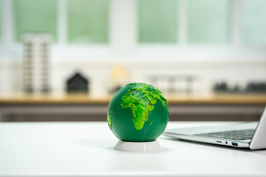 Green globe on a desk beside a laptop with a blurred home office background, symbolizing eco business, sustainability and global technology