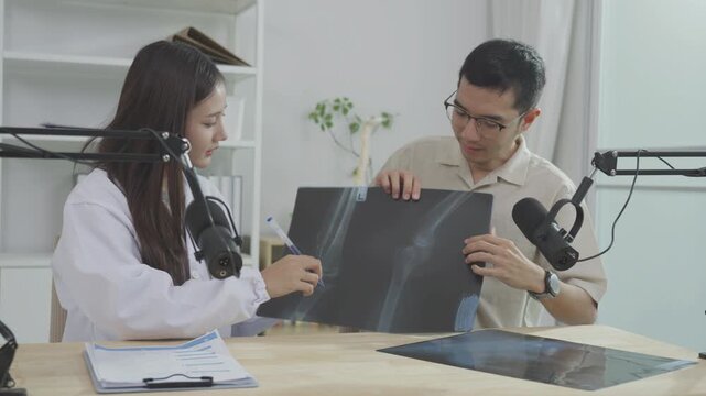 Asian female doctor explaining a chest X-ray film to a male host during a medical podcast interview. Recording a healthcare audio broadcast in a clinic studio. Respiratory health concept.