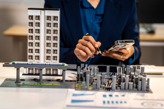 Business person using smartphone and stylus while reviewing an architectural model and city plan on a desk