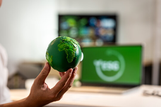 Hand holding a small green earth globe with blurred screens in the background, showing a concept of sustainability, environment, and modern media.