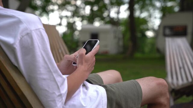 Person lounges in chair, scrolling phone outdoors. Relaxed posture, focused on device. Background shows campsite with trailers. Ideal for vacation, travel, or digital lifestyle themes