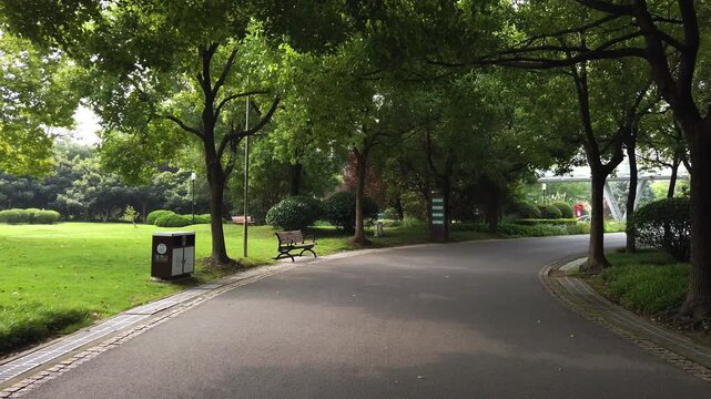 A curving paved path winds through a tree-lined park, with green lawns and a bench under leafy canopies.
