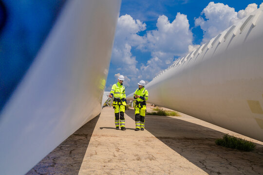 The smooth white upper surface of a stored turbine blade sweeps out of frame toward a blue sky, with two hi-vis workers reduced to bright specks beside its far tip