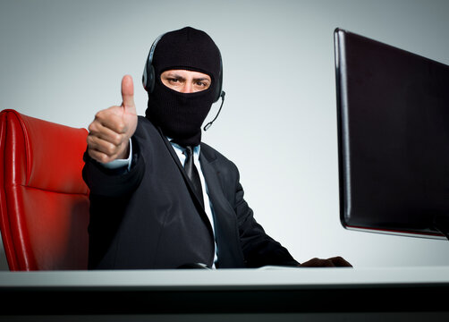 Masked businessman in suit wearing headset showing thumbs up gesture at desk cyber fraud concept isolated on white background.