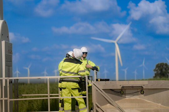 A technician leans forward over the guardrail of a steel platform to look down at the equipment below, with wind turbines standing in the summer fields beyond the barrier