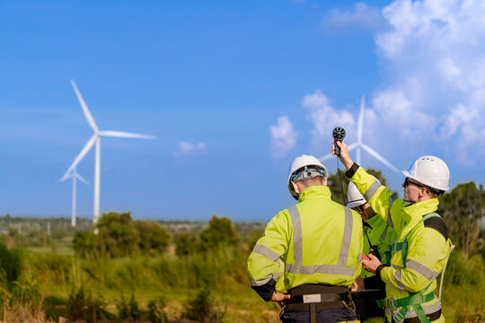Two wind energy workers take a wind speed reading together, one lifting the vane probe skyward while the other stands close behind him in matching yellow hi-vis workwear