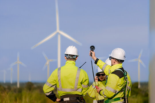 Close view over the shoulders of two technicians as one holds a black vane anemometer out to the side, sunlight rimming their hard hats and reflective yellow collars