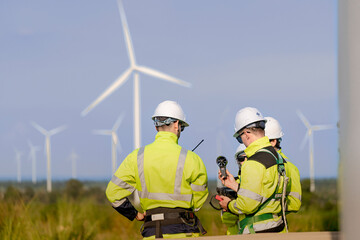 Fototapeta premium A wind technician holds a vane anemometer at chest height while his teammate leans in from the right, both framed against a bright sky with turbines soft in the background