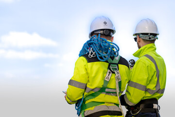 Fototapeta premium Seen from behind, two rope access technicians stand together on the wind farm with a large blue rope coil between them, their white hard hats bright against the pale sky