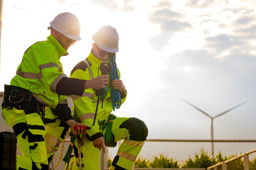 Backlit by the low sun, two wind farm technicians work side by side beside a wire fence, one bending toward a piece of equipment while the other stands close over him © ultramansk