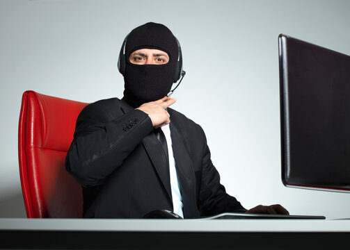 Hacker wearing suit and balaclava mask with headset sitting at desk with computer for cyber crime concept.