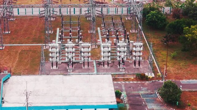 Aerial drone view of a large electrical substation featuring high voltage transformers, transmission towers, steel structures, switchgear, and power distribution equipment surrounded by dense urban bu