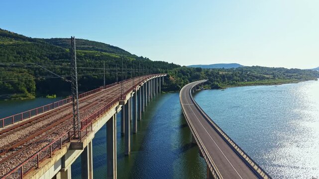 A railway bridge with train tracks stretches over a river while a highway runs parallel to it. The landscape features green hills in the background under a clear sky.