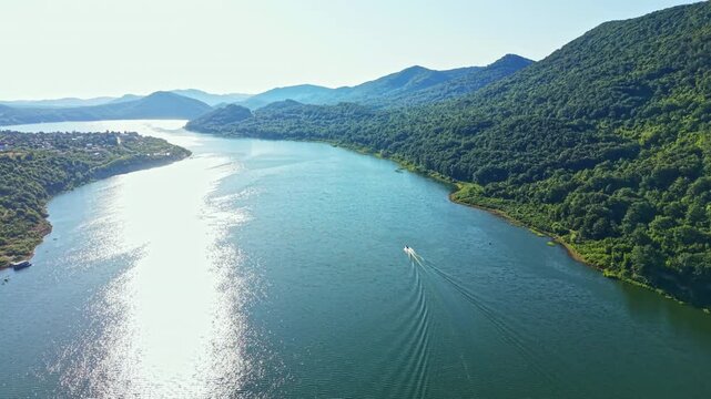 A boat travels along a river with sunlight shining on the water. Green hills border the waterway under a clear sky in the afternoon.