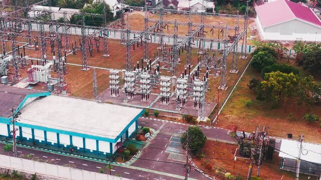 Aerial drone view of a large electrical substation featuring high voltage transformers, transmission towers, steel structures, switchgear, and power distribution equipment surrounded by dense urban bu