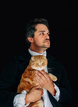 19th century European nobleman in tailcoat holding a red Scottish Fold cat, black background.
