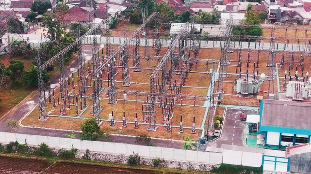 Aerial drone view of a large electrical substation featuring high voltage transformers, transmission towers, steel structures, switchgear, and power distribution equipment surrounded by dense urban bu
