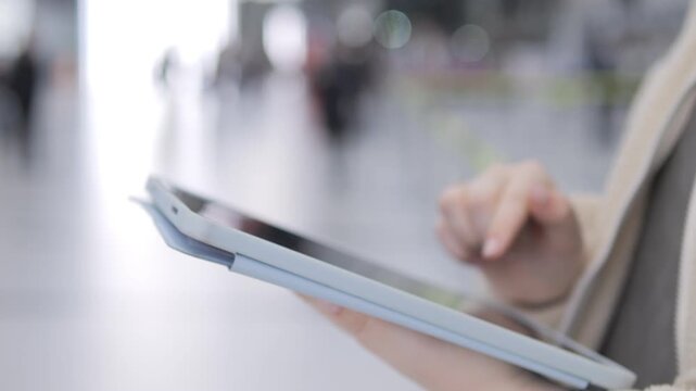 Person holds tablet, taps screen in public space. Background shows blurred people walking. Hand gestures suggest digital interaction. Modern tech used for communication or work