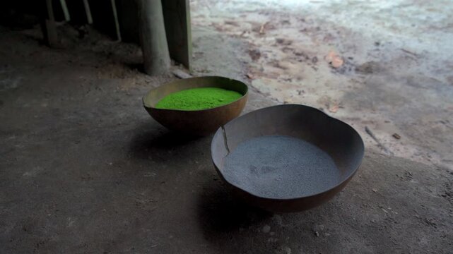 Static medium shot of traditional indigenous mambe green powder and plant ash displayed in gourd bowls on dirt floor inside a maloca in Colombian Amazon.