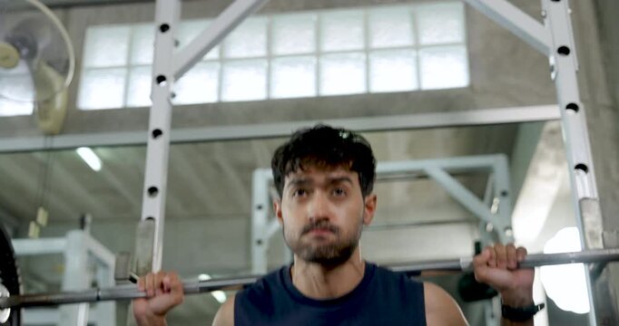 Young Asian man performs barbell shoulder exercise in modern gym, lifting weight from rack with controlled movement, solo upper body strength training, muscle building, wellness, healthy lifestyle.