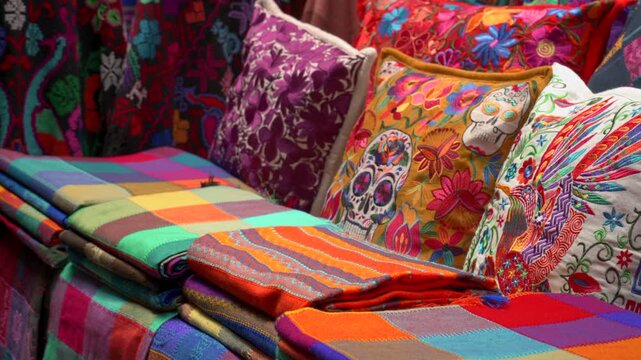Static medium shot of folded colorful Mexican tablecloths and embroidered cushions with Day of the Dead skulls at a souvenir market.