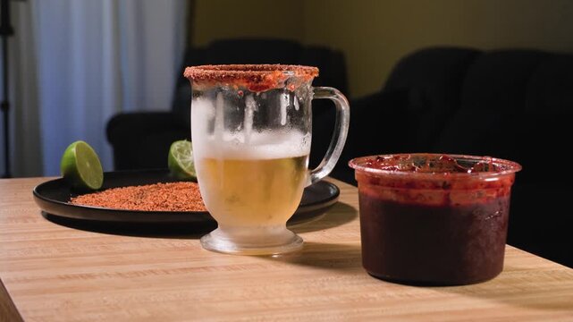 A man returns a frosted plastic cup filled with dark chamoy sauce to a kitchen table beside fresh ingredients