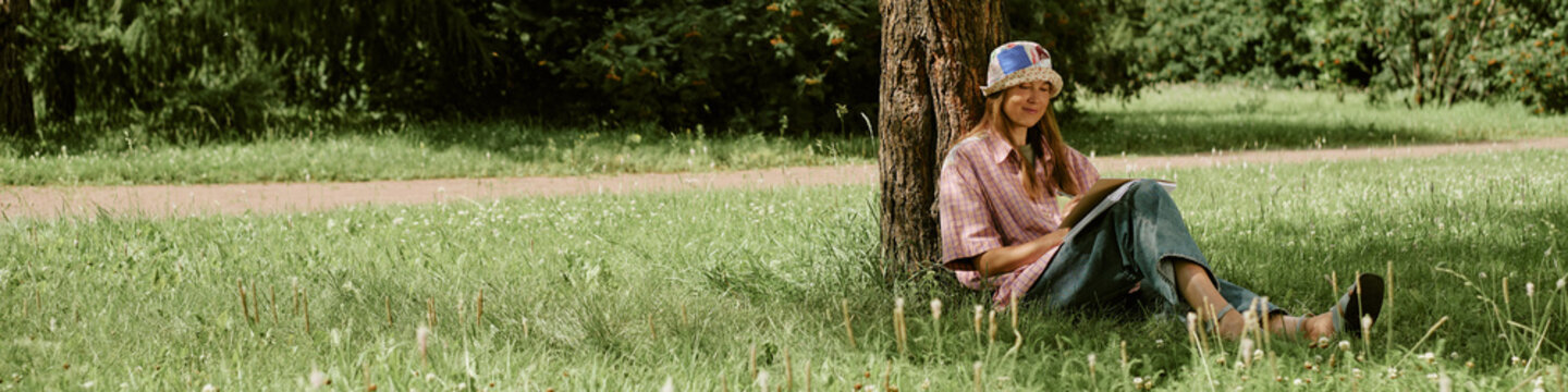 Header of Caucasian young woman sitting on grass under tree drawing in sketchbook during outdoor art master class. Image suitable for promoting creative workshops and leisure activities