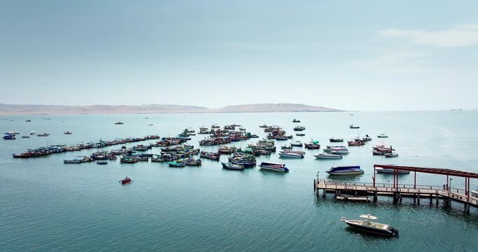 Numerous small fishing and tourist boats anchored in ocean water near pier in Paracas, Peru. Aerial forward, copy space