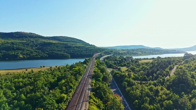 Railroad tracks stretch across a landscape with a river on one side and rolling hills on the other. The sun shines brightly over the scene, showing lush greenery.