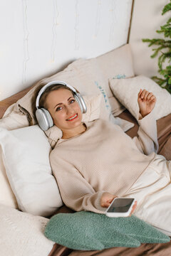 Happy woman lying bed wearing headphones holding phone
