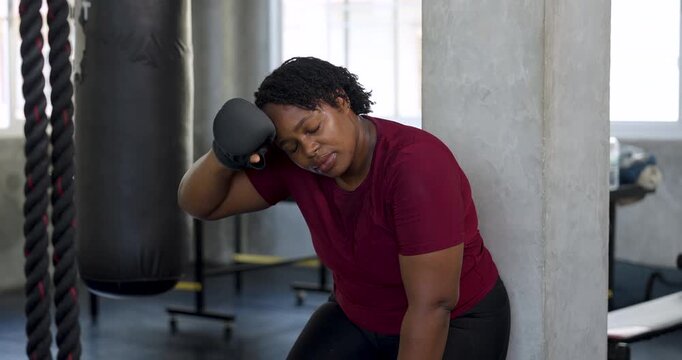 African American plus size woman sits bent forward after boxing workout, resting glove against head in gym. Curvy female athlete cooling down after intense session, wellness recovery, body confidence.