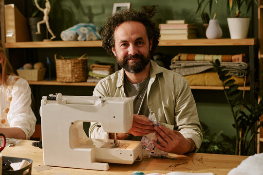 Portrait of middle aged Caucasian man smiling at camera while working with sewing machine in workshop. Image represents creative hobby, craft education, and small business marketing