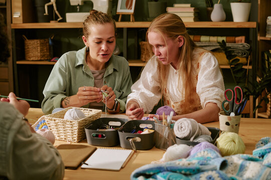 Two Caucasian young adult women learning knitting at workshop table. Image suitable for promoting local craft classes, creative hobbies, and community education programs, medium shot