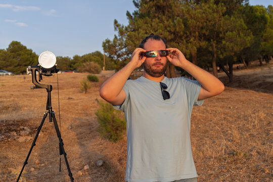 Man observing solar eclipse wearing protective glasses with camera and telescope setup in field at sunset