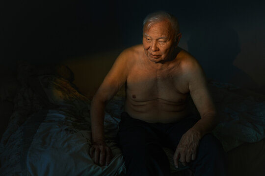 Pensive senior man sits shirtless on a bed at sunset