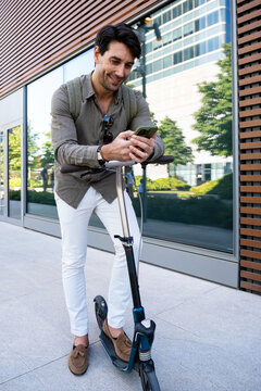 Smiling man uses smartphone while standing with scooter in city