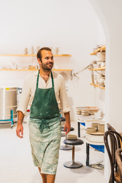 Smiling male ceramist walks in his indoor pottery workshop