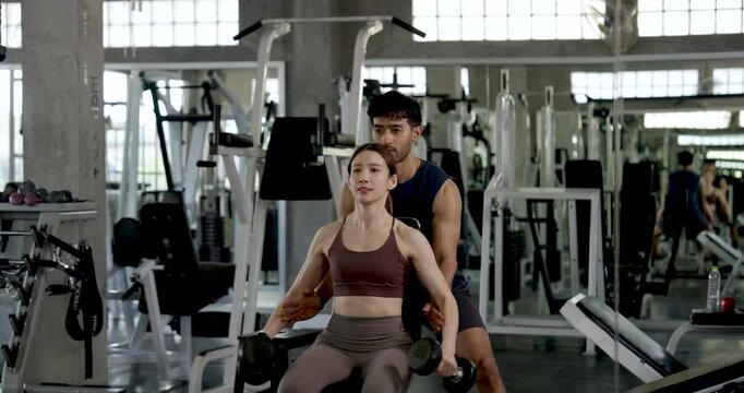 Young Asian couple practicing seated dumbbell lateral raise in modern gym, man spotting woman from behind, partner workout with coaching, arm toning, motivation, teamwork, healthy lifestyle.