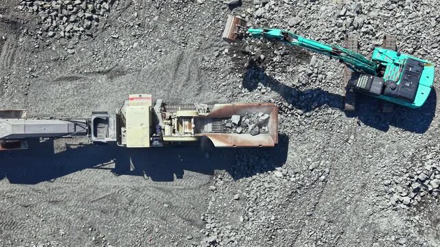 A large machine works in a granite quarry, breaking down rock material and loading it onto a truck. The activity takes place during daylight in a rugged landscape filled with stone.