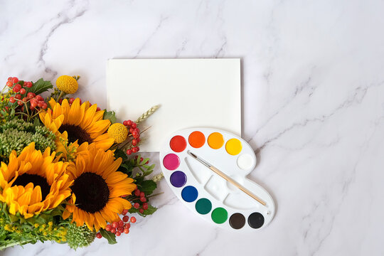 watercolor palette, brush, empty paper sheet and bright autumnal bouquet with sunflowers on marble table background. Desk of creative worker, Painter workplace. symbol of Arts. top view. copy space