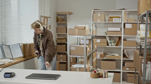 Young adult Caucasian male entrepreneur wearing headphones walking in warehouse, opening laptop to start working. Footage represents small business management and e-commerce logistics
